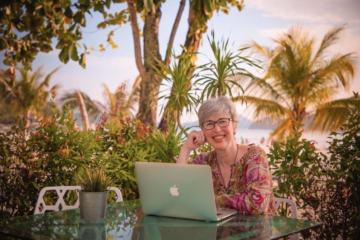 Birgit Arnold sitzt am Laptop auf einer grünen Terrasse am Meer und blickt lächelnd in die Kamera.